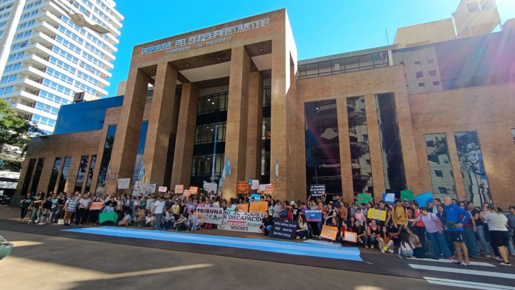 Una multitud se manifiesta frente al edificio de la Cámara de Representantes de Misiones. Los manifestantes sostienen pancartas con mensajes sobre discapacidad y una bandera argentina extendida en el suelo. El edificio de ladrillo visto destaca al fondo bajo un cielo despejado.