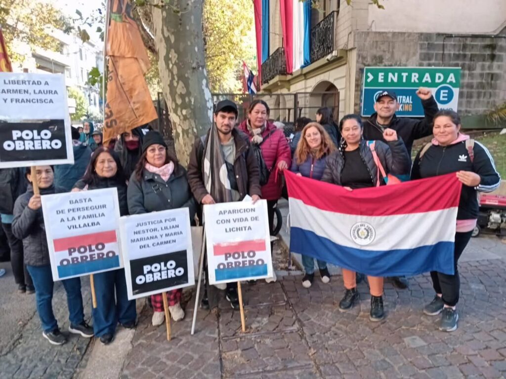 "Compañeres del Polo Obrero en una movilización en la Embajada paraguaya. Los mismos sostienen una bandera de Paraguay (lineas horizontales rojo, blanco y azul) y carteles que dicen Libertad a Carmen, Laura y Francisca; Basta de pereguir a la familia Villalba; Justicia por Nestor, Liliam Mariana y María del Carmen; Aparición con vida de Lichita"