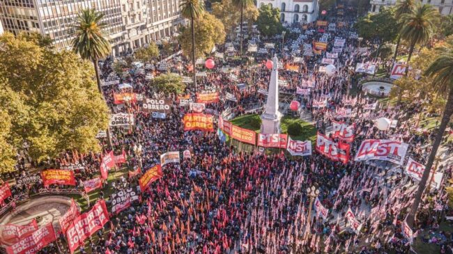 Fotografía aérea de un acto masivo del Frente de Izquierda en Plaza de Mayo, Buenos Aires. La plaza está colmada de personas con banderas rojas. Se destacan carteles del 'Frente de Izquierda de los Trabajadores' y del 'Partido Obrero'. Hay globos rojos de gran tamaño flotando sobre la multitud y edificios históricos al fondo, como la Catedral Metropolitana.