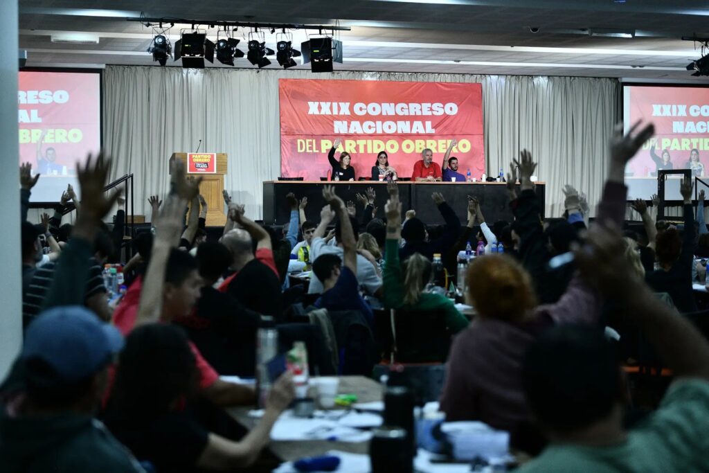 "Vista desde el fondo de un salón lleno de gente donde muchas personas tienen las manos levantadas para votar o participar. Al frente, un panel de cuatro personas está sentado en una mesa larga sobre un escenario. Detrás de ellos hay una gran pancarta roja que dice 'XXIX CONGRESO NACIONAL DEL PARTIDO OBRERO' en letras blancas. Hay pantallas a los lados mostrando lo mismo y un podio con un logo de 'PARTIDO OBRERO' a la izquierda."