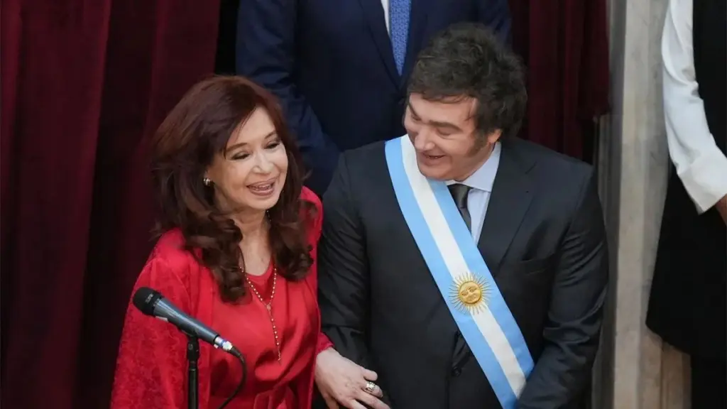 "Fotografía de Cristina Fernández de Kirchner y Javier Milei compartiendo un momento de risas durante la ceremonia de asunción presidencial en el Congreso de la Nación Argentina. Ella viste un conjunto de color rojo intenso y él lleva la banda presidencial celeste y blanca sobre un traje oscuro. A la izquierda se observa parcialmente a un granadero con uniforme de gala."