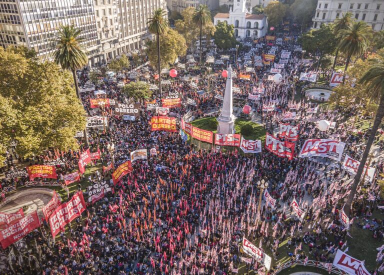 Fotografía aérea de una manifestación masiva en Plaza de Mayo, Buenos Aires. La plaza está colmada de personas con banderas rojas. Se destacan carteles del 'Frente de Izquierda de los Trabajadores' y del 'Partido Obrero'. Hay globos rojos de gran tamaño flotando sobre la multitud y edificios históricos al fondo, como la Catedral Metropolitana.