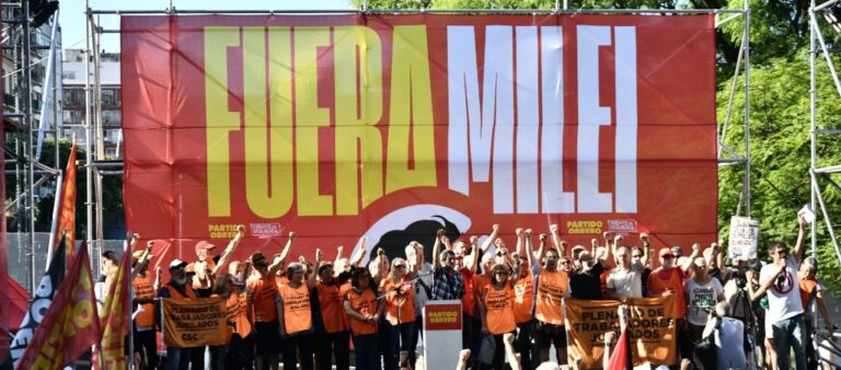 Fotografía de un acto político en un escenario al aire libre. Al fondo, un gran telón rojo muestra la consigna 'FUERA MILEI' en letras grandes de color amarillo y blanco. Sobre el escenario, un grupo de aproximadamente veinte personas posa con los puños en alto. La mayoría viste pecheras de color naranja. Frente al escenario, una multitud de espaldas observa el acto, con banderas rojas visibles en los laterales.