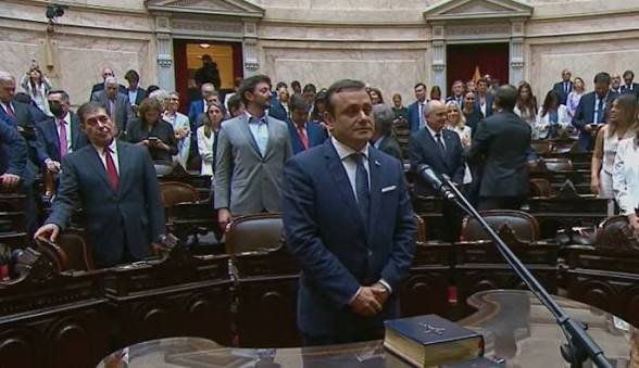 Oscar Herrera Ahuad de pie frente al estrado en la Cámara de Diputados, jurando como Diputado Nacional por la provincia de Misiones. Viste traje azul, tiene las manos entrelazadas y está rodeado de legisladores en sus bancas durante una sesión oficial.