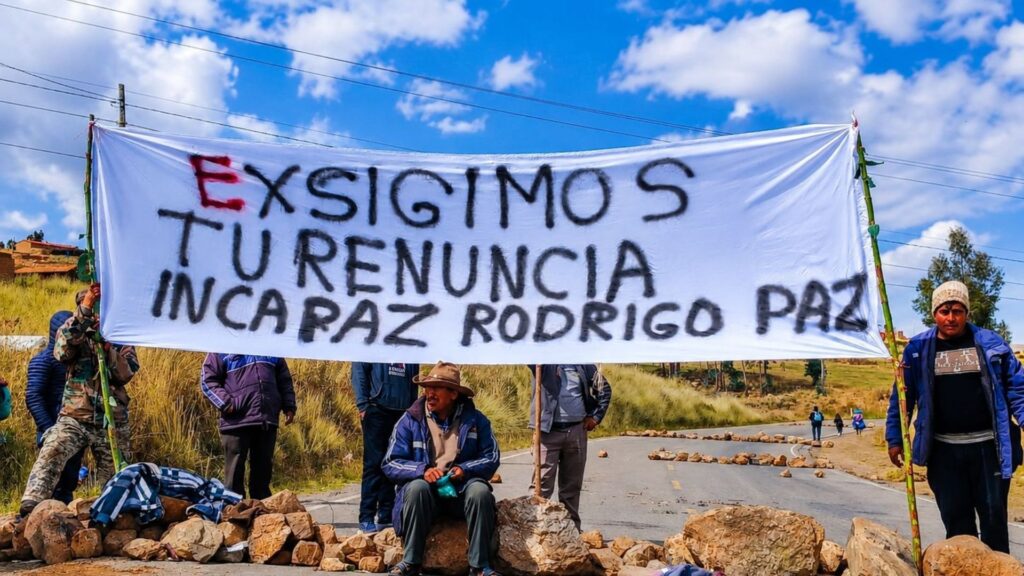 "Un grupo de campesinos que ha bloqueado una ruta rural en Bolivia con grandes rocas en un paisaje montañoso. Sostienen una pancarta blanca pintada a mano con un mensaje en español en tres líneas, que dice: 'EXSIGIMOS TU RENUNCIA INCAPAZ RODRIGO PAZ', con algunas letras en rojo. Un manifestante está sentado sobre las rocas en primer plano, y otros están de pie detrás de la pancarta. El fondo muestra colinas y un cielo azul con nubes blancas."
