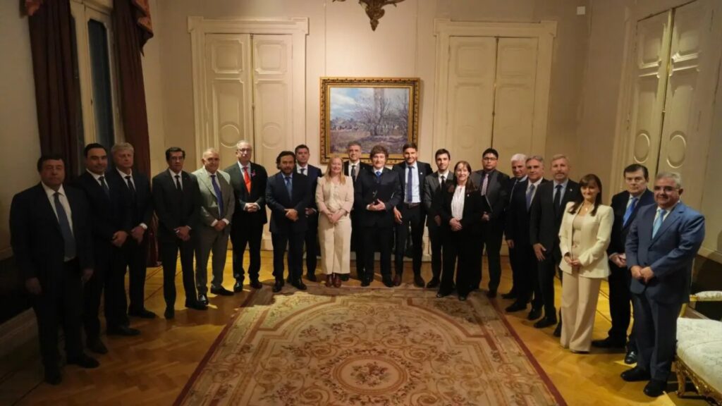 "Fotografía grupal de Javier Milei junto a gobernadores y funcionarios en un salón formal de la Casa Rosada. El grupo de 23 personas está dispuesto en forma de 'U' sobre una alfombra ornamental. En el centro, Milei sostiene una pequeña estatuilla. El ambiente cuenta con paredes color crema, puertas de madera tallada de doble hoja y un cuadro central. La iluminación es cálida y la atmósfera institucional."
