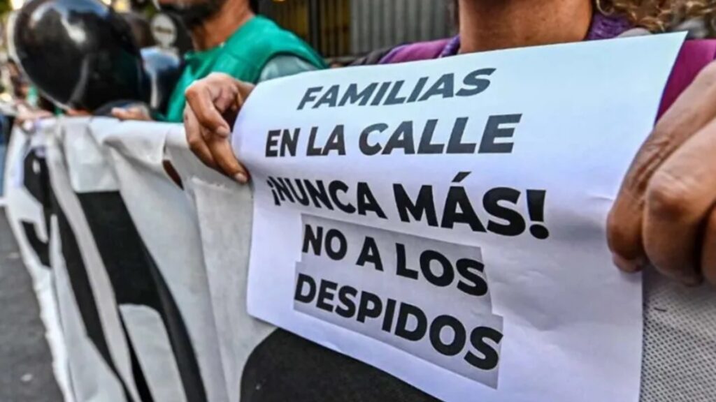 "Primer plano de una persona sosteniendo un cartel blanco impreso en español durante una manifestación callejera. El texto dice: 'FAMILIAS EN LA CALLE ¡NUNCA MÁS! NO A LOS DESPIDOS'. Las manos de la persona sujetan el cartel por ambos lados. Al fondo, borrosos, se distinguen otros manifestantes y el contorno de un casco de motocicleta."