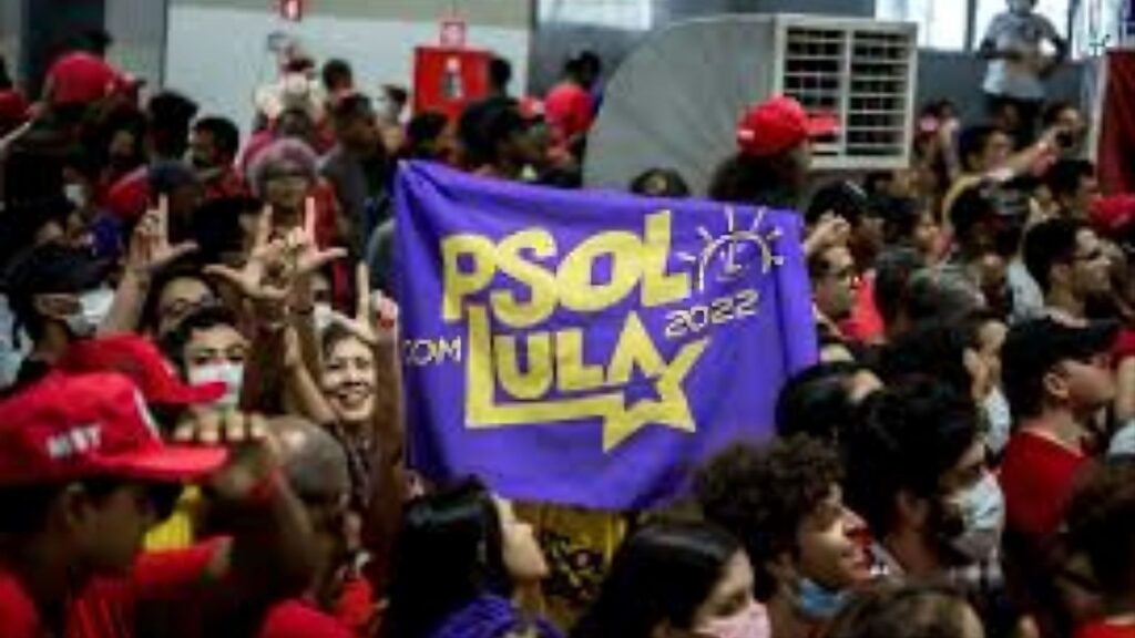 "Una multitud en un acto político en Brasil. En el centro, una bandera violeta con letras amarillas dice 'PSOL com LULA 2022'. A la izquierda, varias personas realizan con sus manos la seña de la letra 'L' y otras señas de apoyo. Predominan las gorras y vestimentas rojas en el resto de la concurrencia."