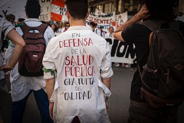 "Un trabajador de salud pública con su uniforme puesto en una movilización. Se encuentra de espaldas, donde se encuentra escrito 'en defensa de la salud publica, gratuita, de calidad' de fondo hay mas personas y pancartas"