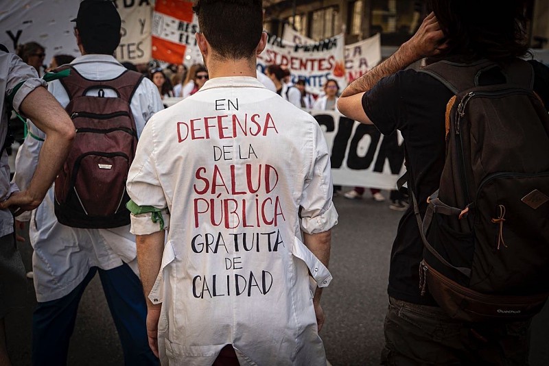 "Un trabajador de salud pública con su uniforme puesto en una movilización. Se encuentra de espaldas, donde se encuentra escrito 'en defensa de la salud publica, gratuita, de calidad' de fondo hay mas personas y pancartas"